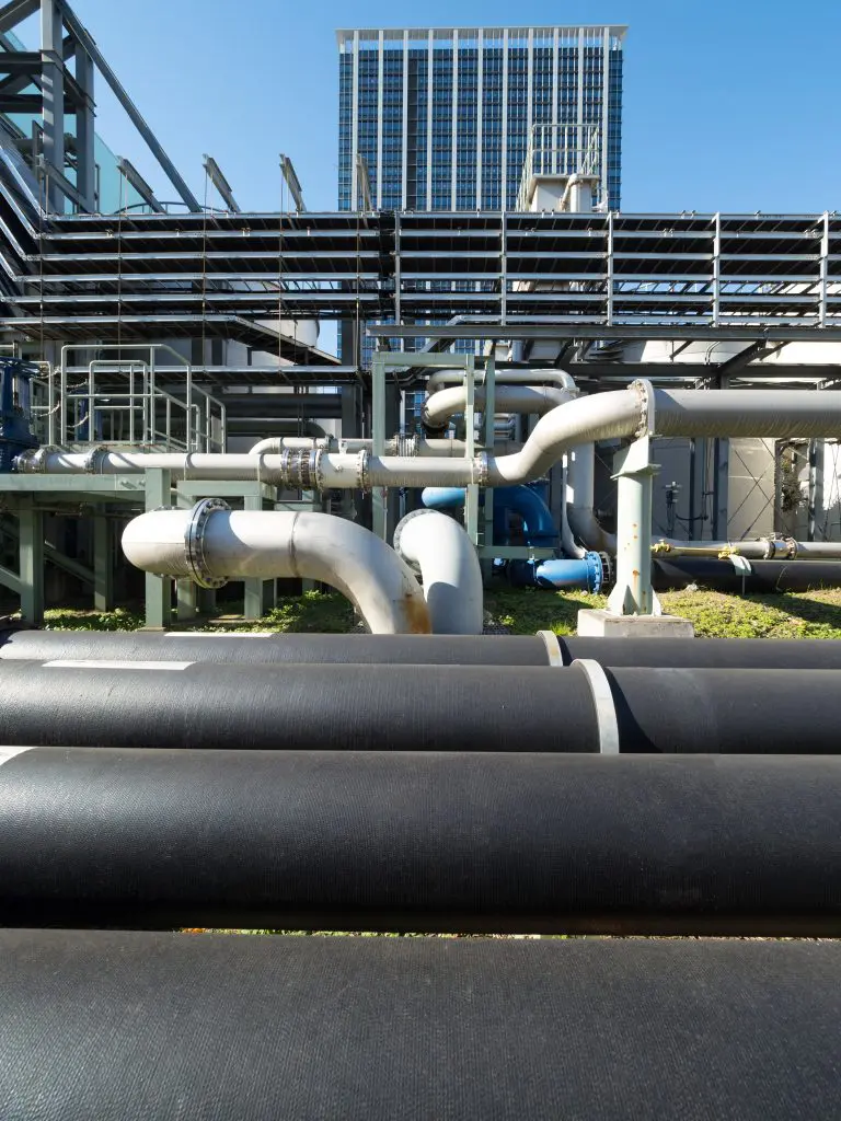 Large black insulated pipes in the foreground of an outdoor mechanical plant, with metal-jacketed piping, steel framework, and a high-rise building under a clear blue sky